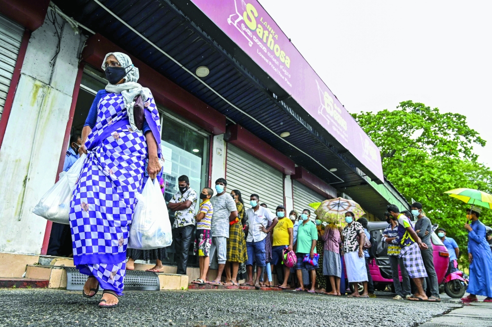 A woman carrying food bags walks pasts people standing in queue outside a state-run supermarket to buy essential food items in Colombo on Friday. -- AFP
