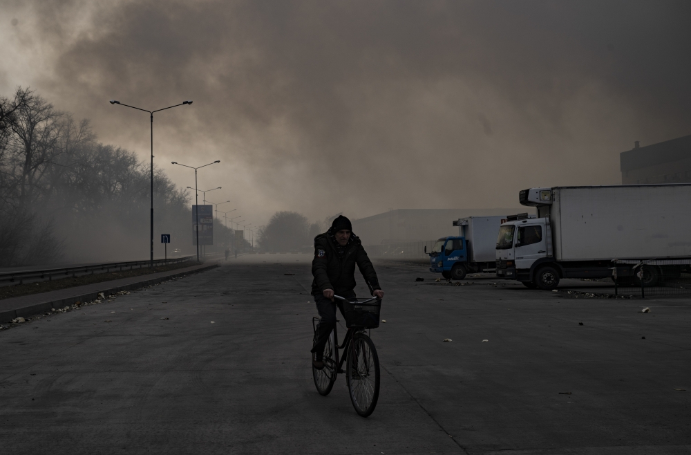 A man cycles through the smoke billowing from a frozen foods warehouse that was hit by artillery fire in Brovary, east of Kyiv, on Saturday morning, March 12, 2022. (Lynsey Addario/The New York Times)
