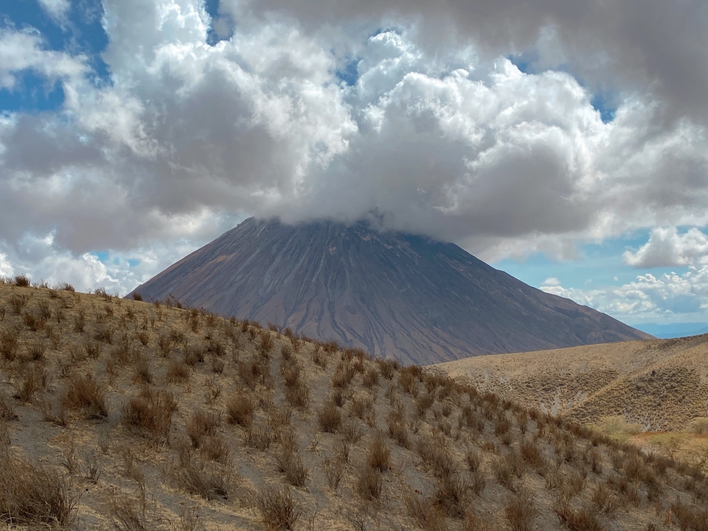 The cloud-topped summit of Ol Doinyo Lengai, in Tanzania on Nov. 11, 2022, an active volcano that last exploded in 2008, looms over the crater highlands, with the outlines of hardened lava flows striping the slopes. (Patrick Scott/The New York Times)