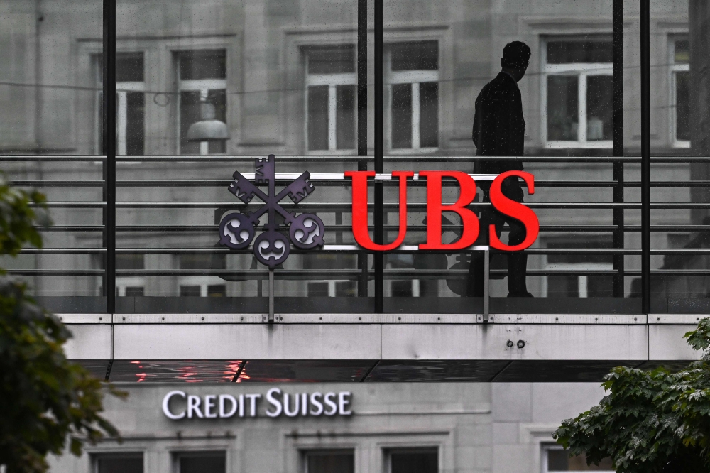 An employee of multinational investment bank UBS walks past a sign of Swiss giant banking UBS above a sign of Credit Suisse in Zurich