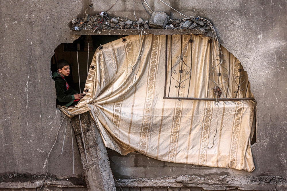 A boy sits by a hole in a wall covered up with canvas in a building overlooking another destroyed by Israeli bombardment in Rafah. — AFP 