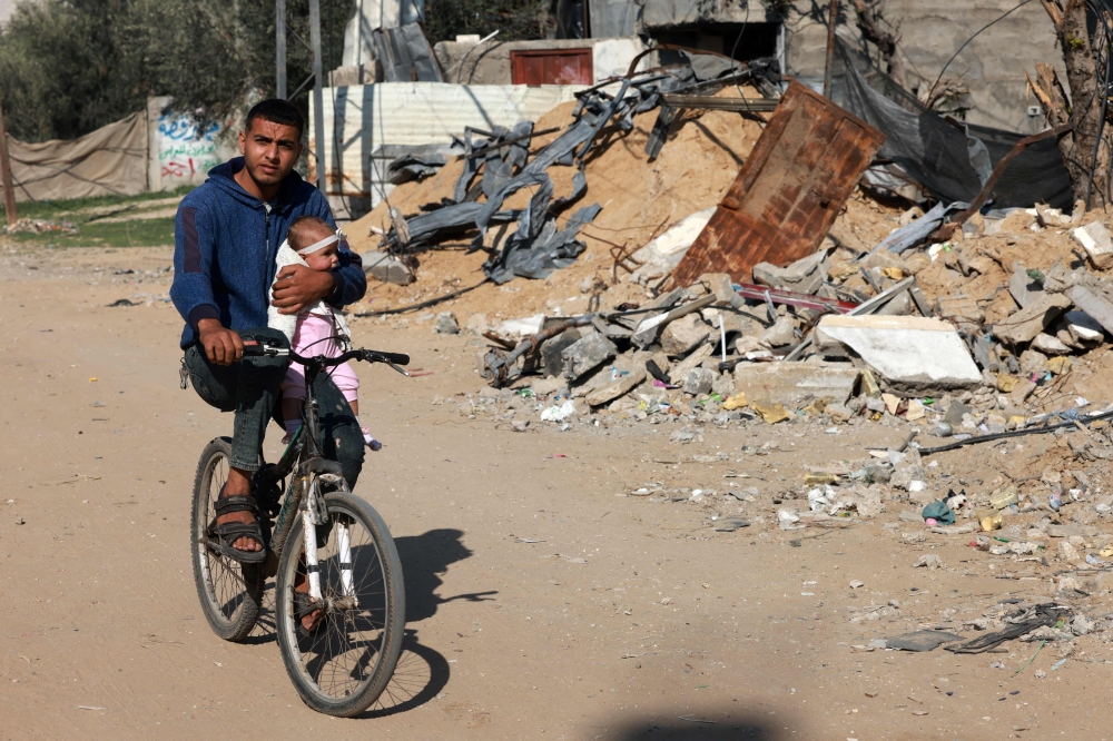 A man carries a baby as he rides a bicycle past a building destroyed during Israeli bombing in Rafah in the southern of Gaza Strip, on December 31, 2023, amid the ongoing conflict between Israel and the Palestinian militant group Hamas. 
