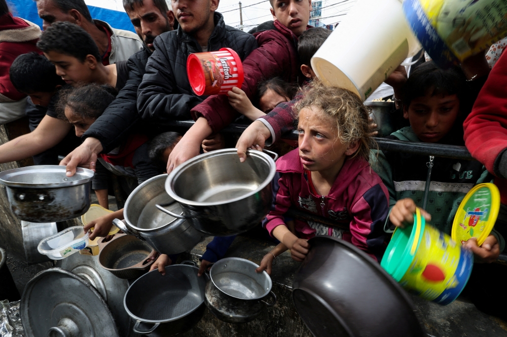 Palestinians with children wait to receive food cooked by a charity kitchen amid shortages of food supplies