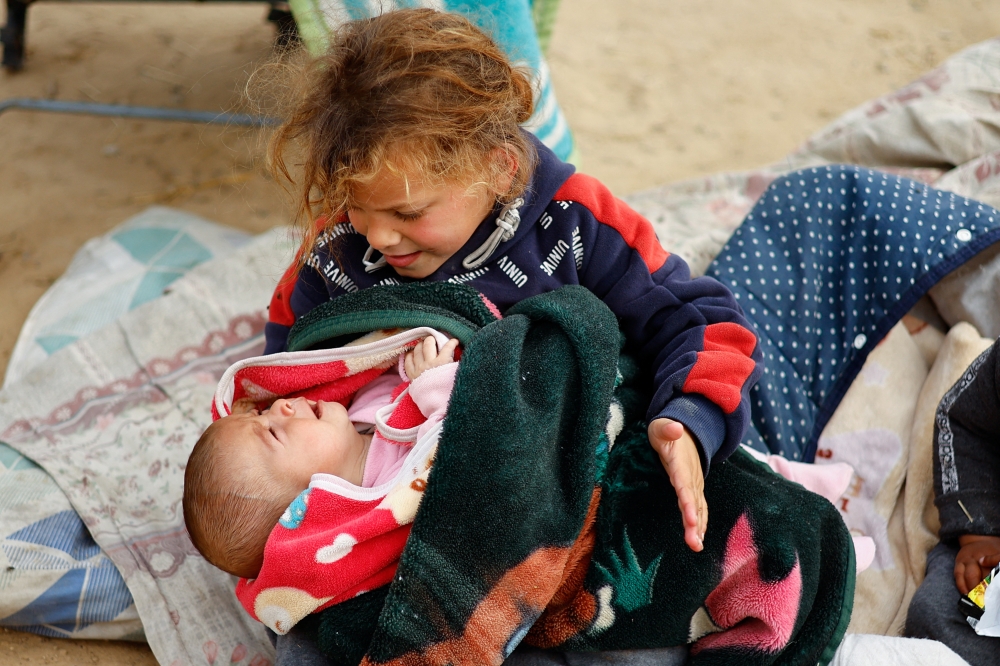 A displaced Palestinian child who fled her house due to Israeli strikes holds a crying baby