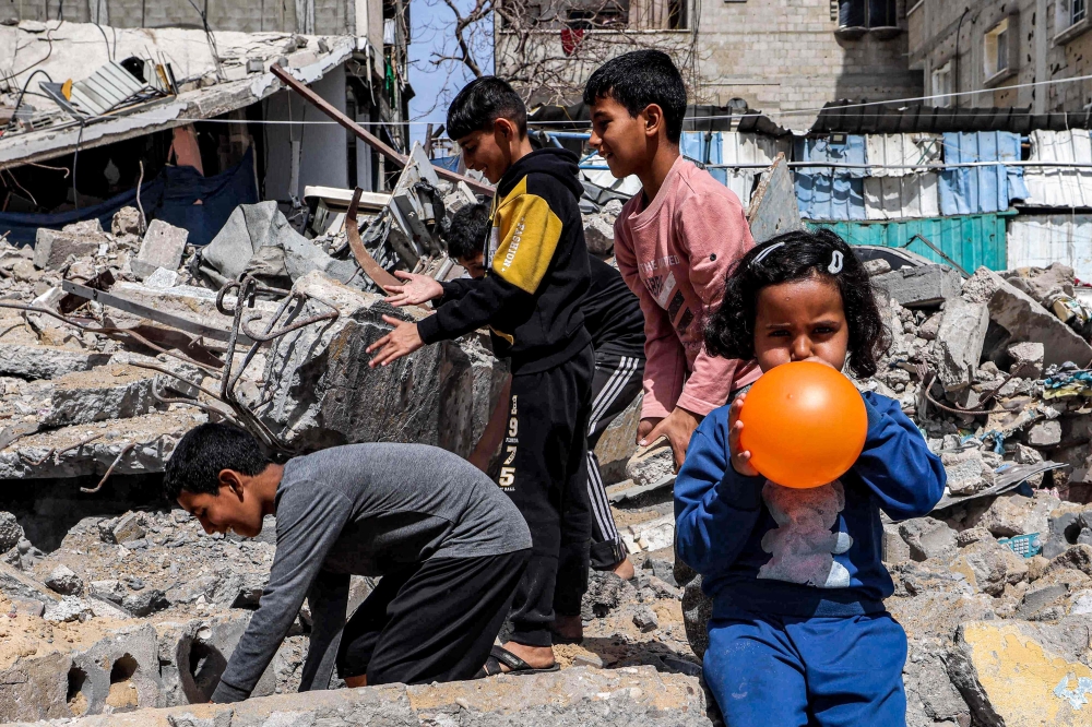 A girl blows a balloon as behind her boys search through the rubble of a destroyed building in Rafah in the southern Gaza Strip amid the ongoing conflict in the Palestinian territory between Israel and Hamas