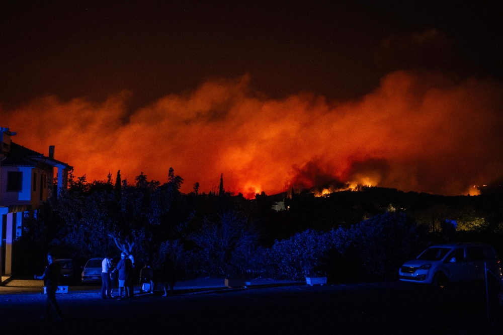 Local residents stand along a street as a wildfire burns in a hillside in Varnavas, north of Athens