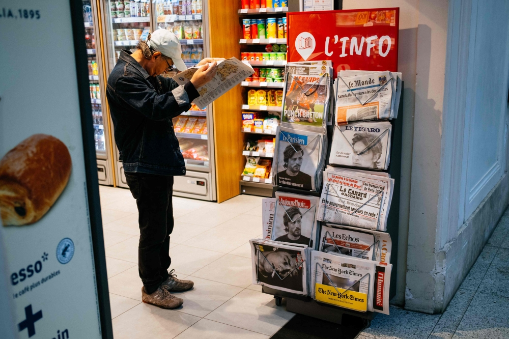 A man reads a newspaper as he stands in a newsagent's booth announcing the death of French actor Alain Delon