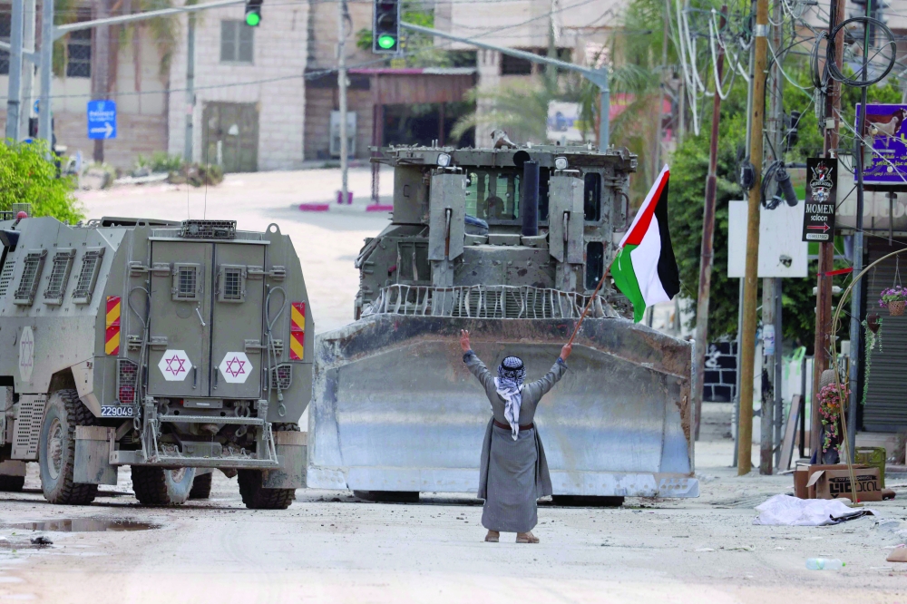 A Palestinian man lifts a national flag and flashes the victory sign as Israeli armoured vehicles drive on a street during a raid in Tulkarem on Tuesday. - AFP