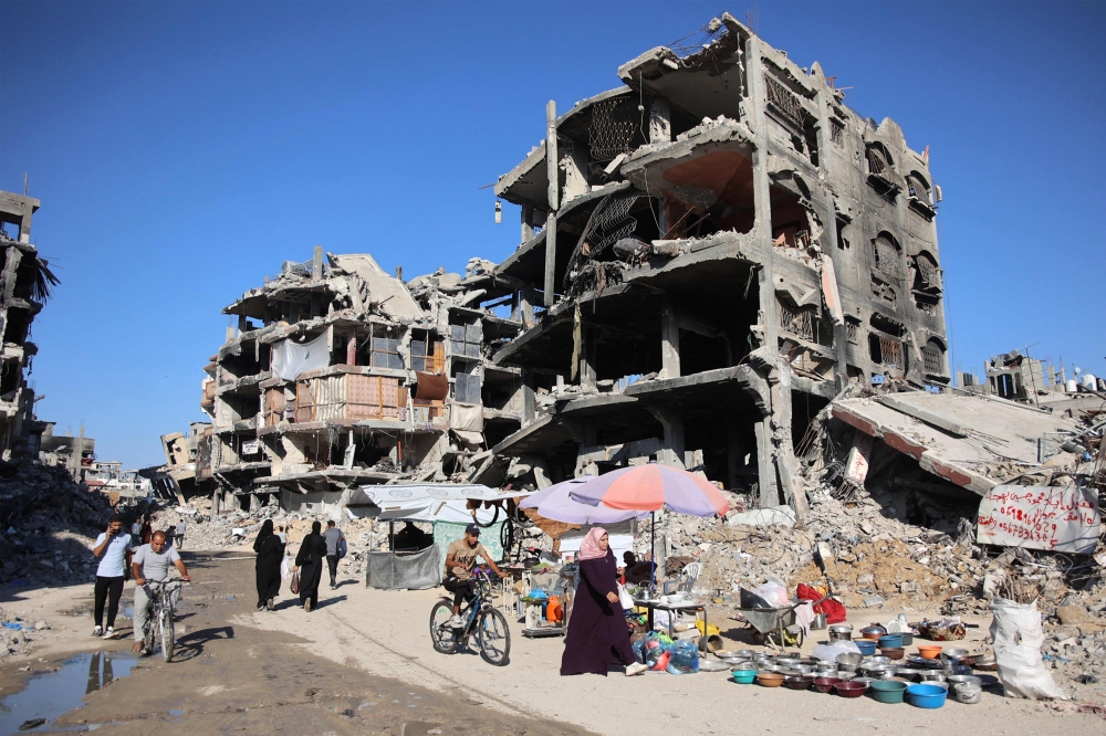 A man cycles past vendors displaying wares outside heavily damaged buildings in a camp sheltering displaced people in Jabalia. — AFP 