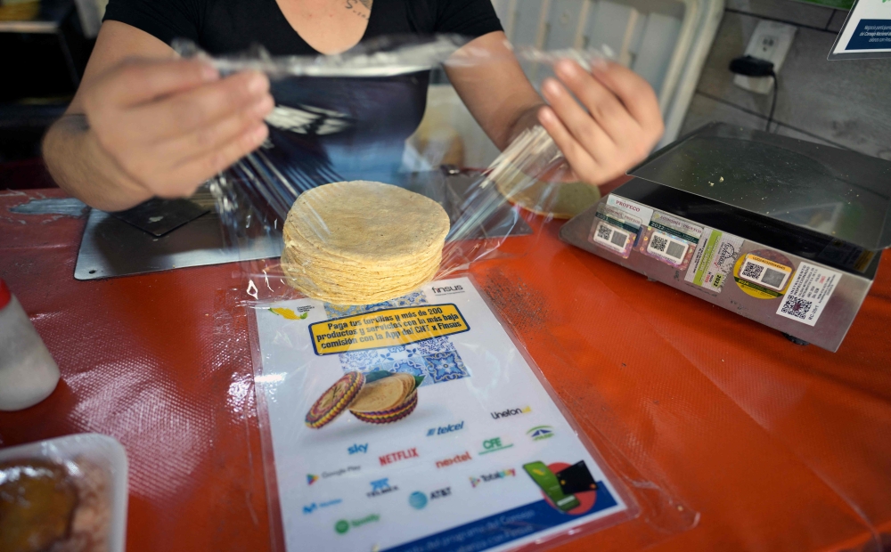 A worker handles tortillas over a sign with information about an app to buy tortillas at a tortilla factory in Mexico City