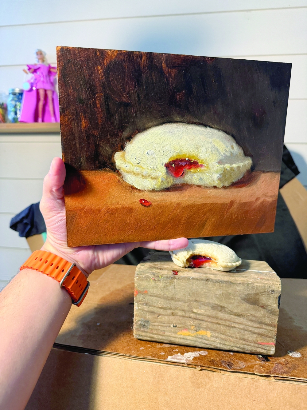 Noah Verrier’s hand holds up a painting of an Uncrustables sandwich above the sandwich that inspired it. 