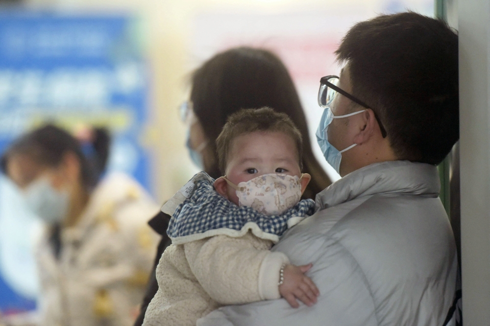 A baby wearing a face mask waits with family members 