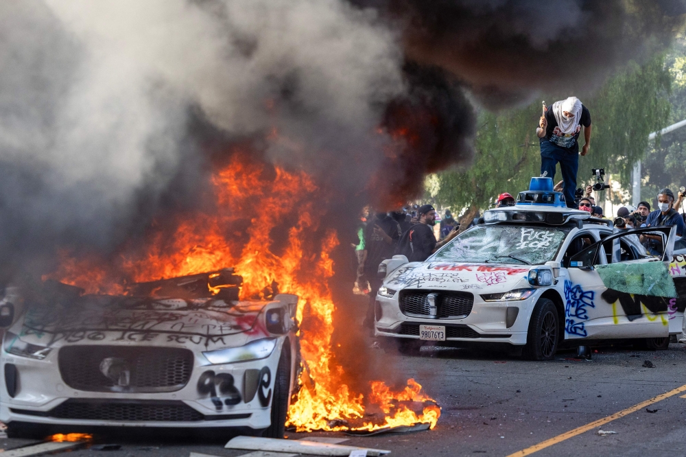 Demonstrators smash the windshield of a vehicle next to a burning Waymo vehicle as protesters clash with law enforcement in the streets surrounding the federal building during a protest following federal immigration operations in Los Angeles, California, on June 8, 2025.