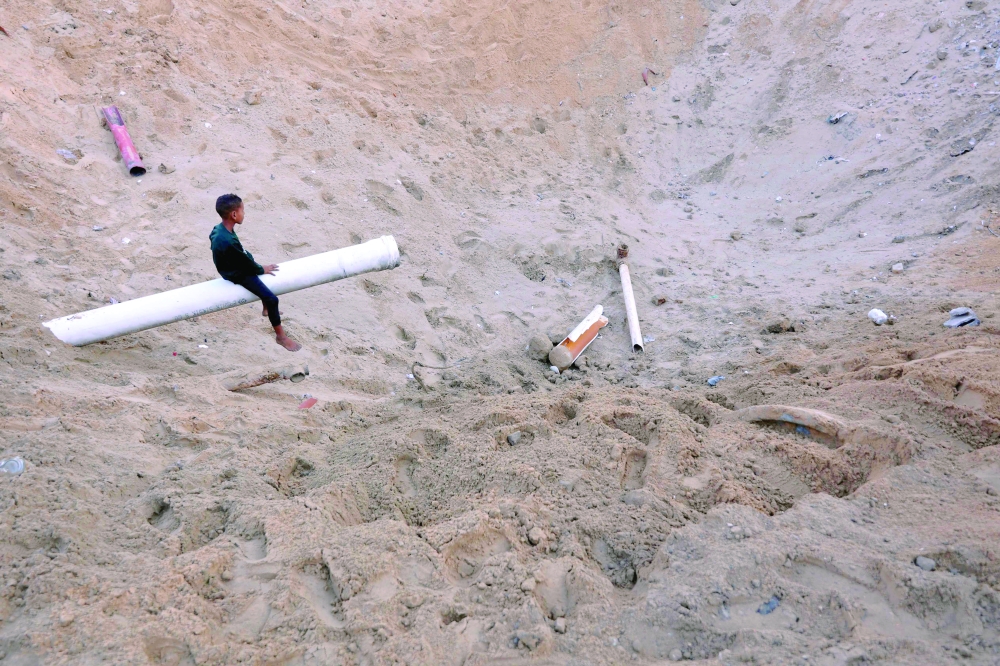 A child sits on what remains of a water pipe in a huge crater after the Israeli army targeted the tents of displaced people in Al Rimal on Saturday. — AFP