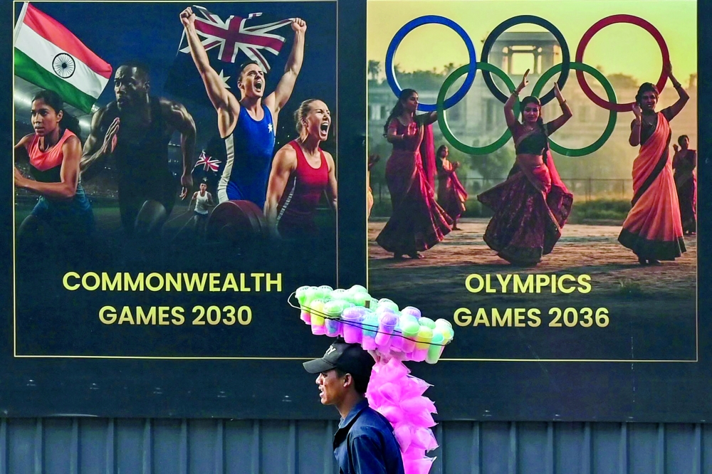 A cotton candy seller walks past a billboard featuring potential sporting events in Ahmedabad on November 26, 2025. The Indian city of Ahmedabad will stage the 2030 multi-sport Commonwealth Games, it was confirmed on November 26. The choice of Ahmedabad, also known as Amdavad, was ratified by a decision of the Commonwealth Sport General Assembly in Glasgow following a recommendation from the executive board. (Photo by Shammi MEHRA / AFP)