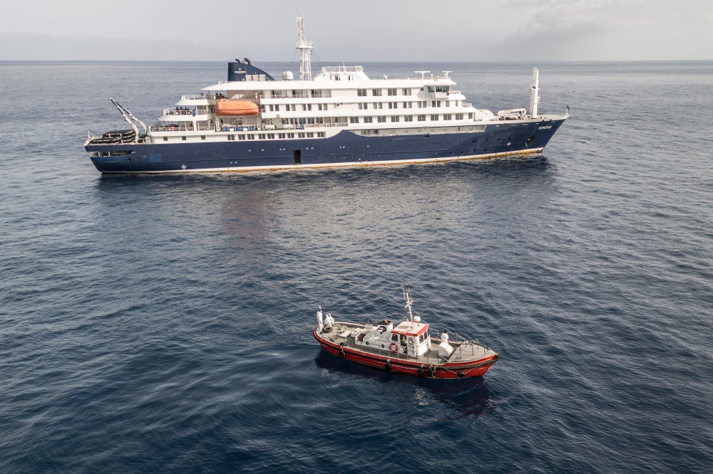 An aerial view of an ambulance boat carrying crew members wearing hazmat suits as they approach the pilot door on the starboard side of the cruise ship MV Hondius, while stationary off the port of Praia, the capital of Cape Verde, on May 5, 2026. 