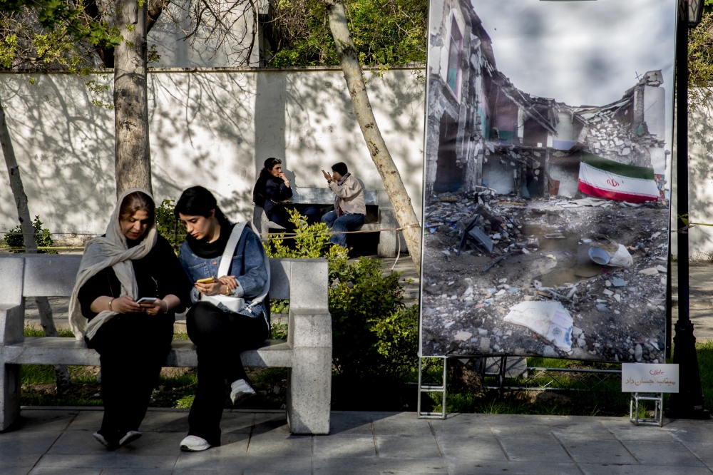 Two women watch their phones next to banner showing destroyed building, in Tehran. — NYT file photo
