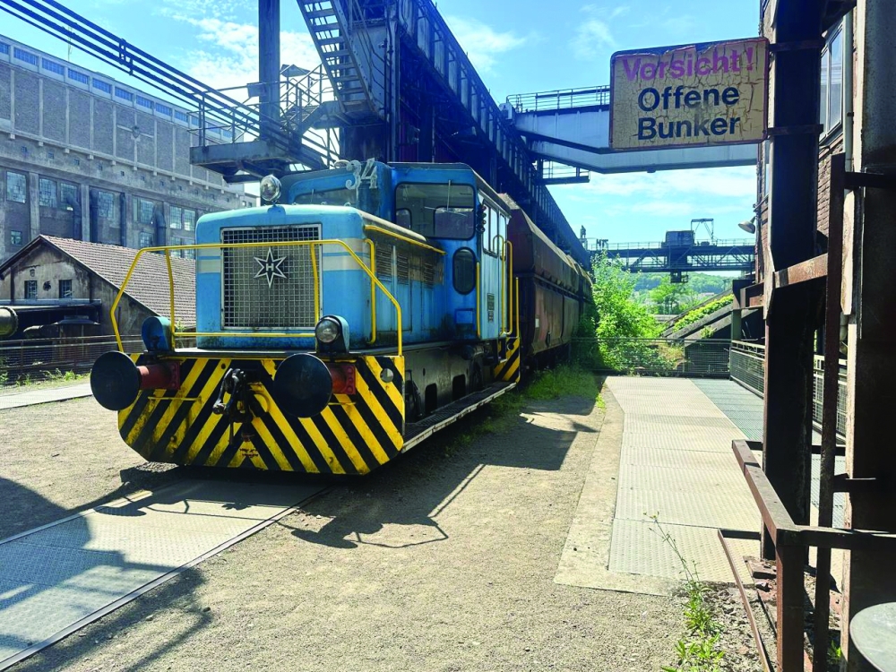 A classic blue and yellow locomotive at the historic Völklingen Ironworks, with the warning sign visible overhead.
