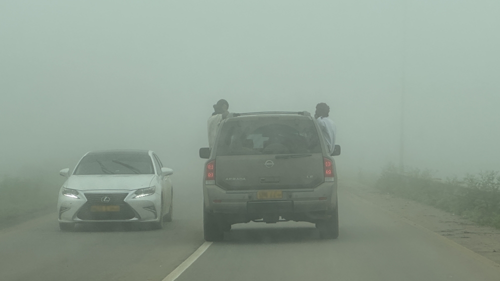 Two passengers lean partly outside a moving vehicle as dense Khareef fog restricts visibility on a road in Dhofar.