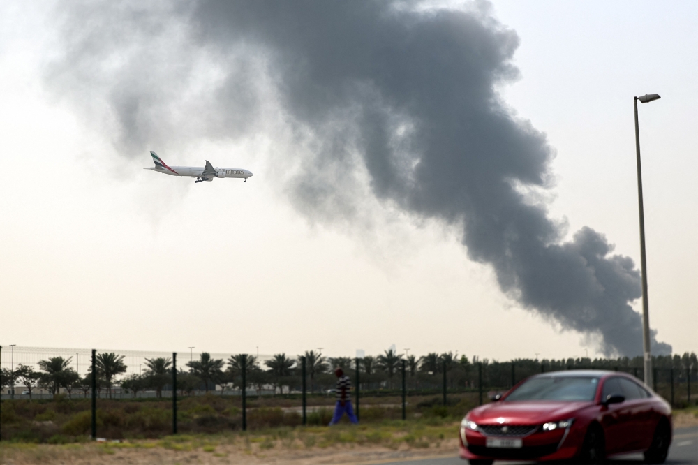 An aircraft prepares for landing as a smoke plume rises from a fire near Dubai Airport in Dubai on March 16, 2026