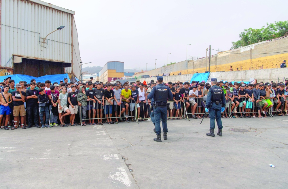 Spain's National police officers stand as migrants wait during a food distribution at the El Tajaral industrial site of Ceuta. — AFP