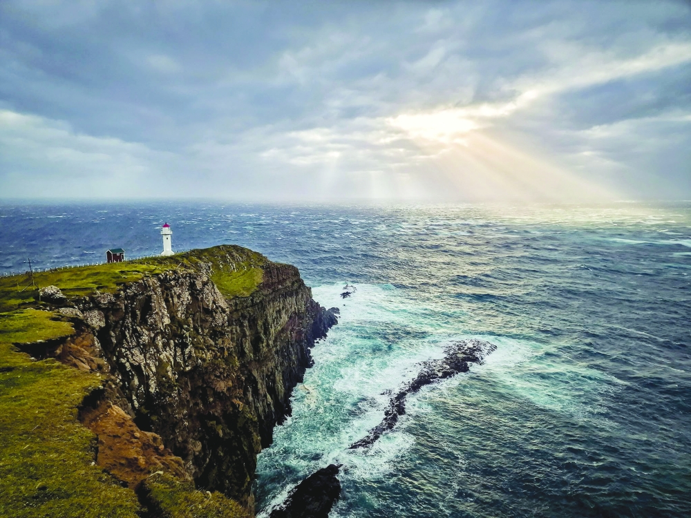 Dramatic coastal scenery in the Faroe Islands, featuring a solitary lighthouse overlooking turbulent seas under rays of sunlight.