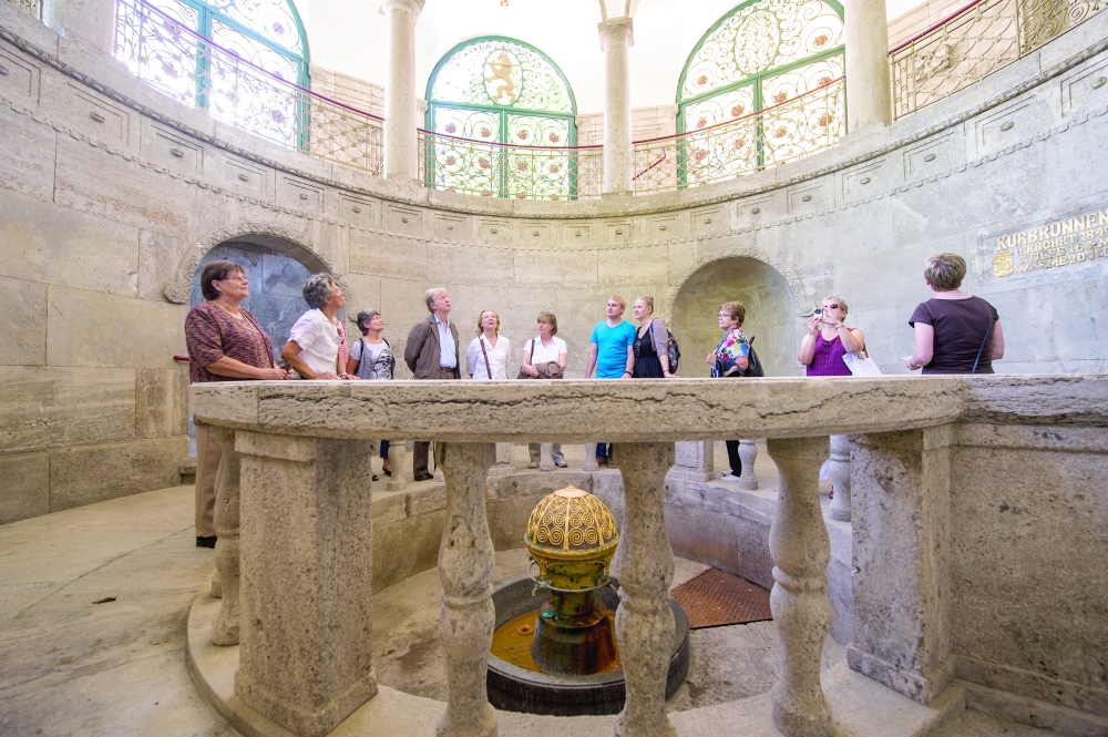 Visitors gather around the historic spring with its ornate golden cover during a guided tour of Bad Nauheim’s Trinkkuranlage.