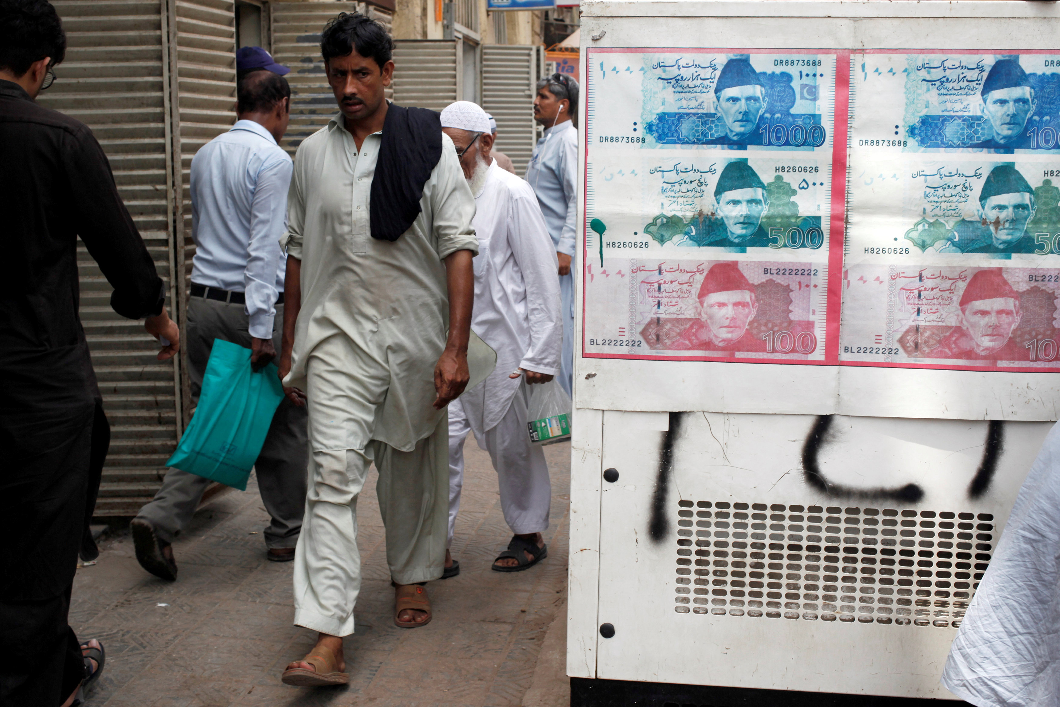 Passersby walk past an advertisement board with photos of Pakistani rupee at a money exchange along a sidewalk in Karachi