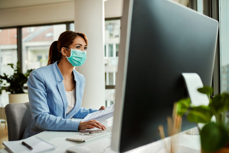 Businesswoman with face mask using computer while working on reports in the office.